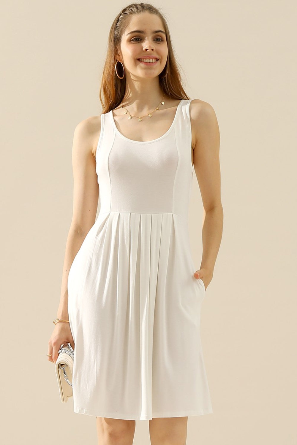 A woman in the Doublju Full Size Round Neck Ruched Sleeveless Dress with Pockets, featuring a stretchy gray fabric, smiles while holding a quilted white purse. She is wearing a gold necklace and bracelet.