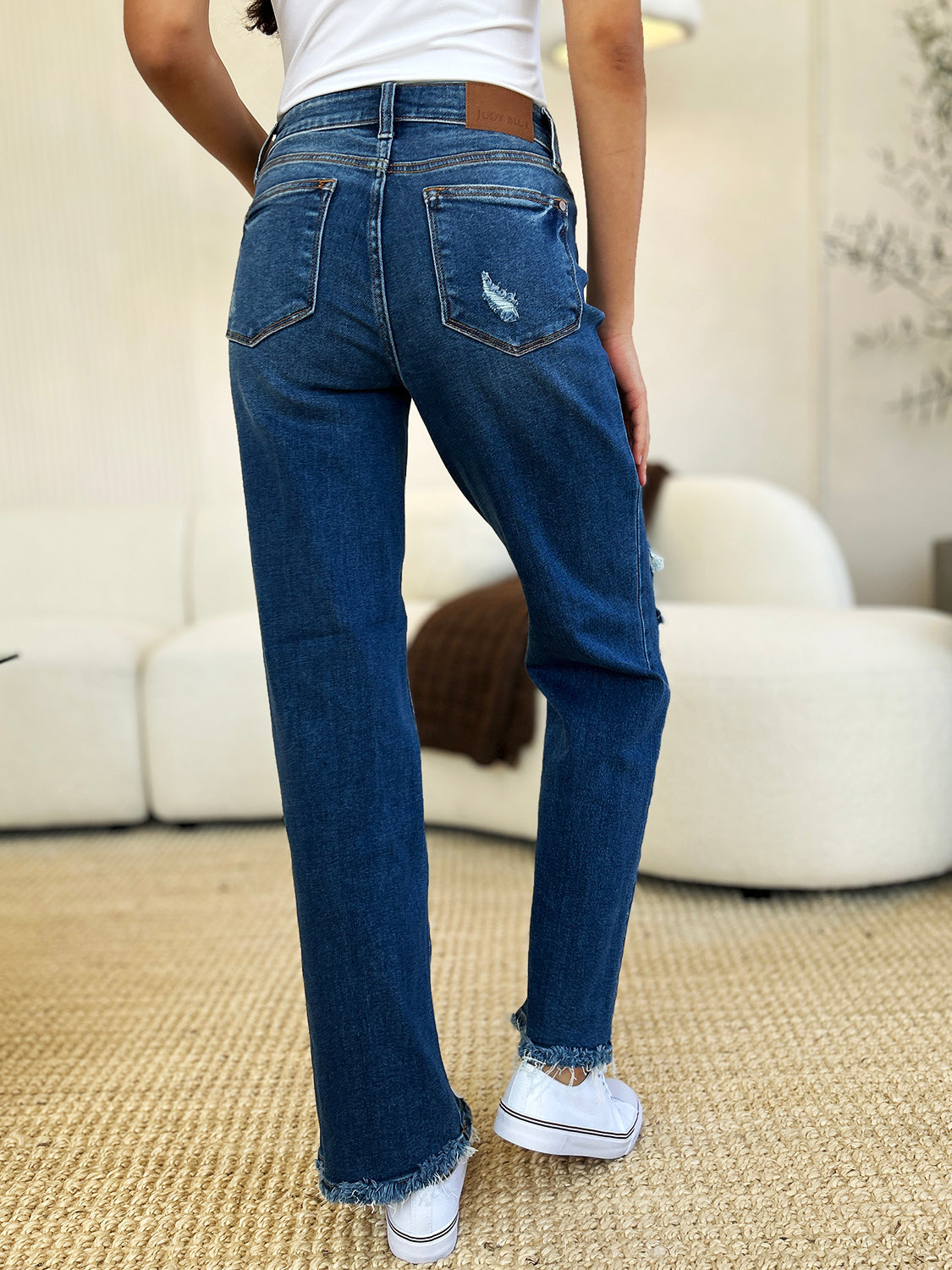 Person standing indoors wearing Judy Blue Full Size Mid Rise Distressed Raw Hem Jeans and black sneakers.