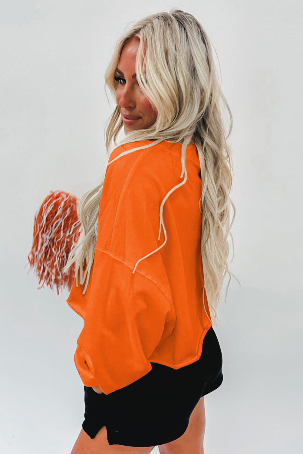 A woman with blonde hair, wearing an Orange Game Day Lettering Rugby Football Notched Neck Sweatshirt and blue jeans, stands next to a decorated Christmas tree.