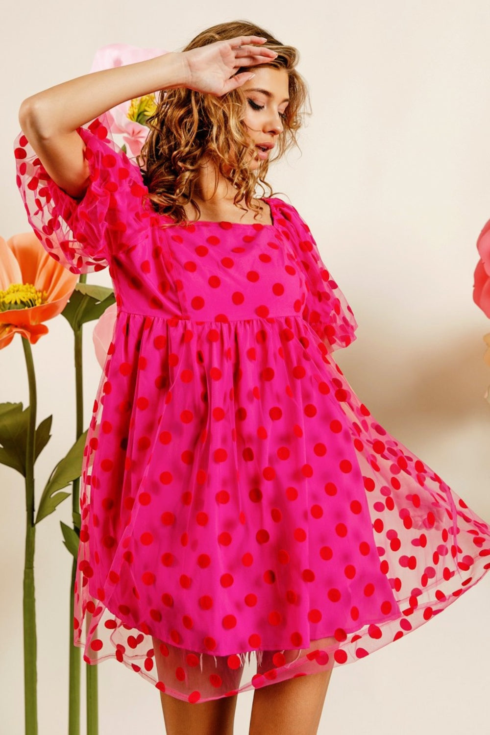 Wearing the BiBi Polka Dot Mesh Puff Sleeve Dress, a woman poses gracefully with one hand on her head. The vintage-inspired pink dress adorned with red polka dots enhances her elegance as she stands amidst large artificial flowers.