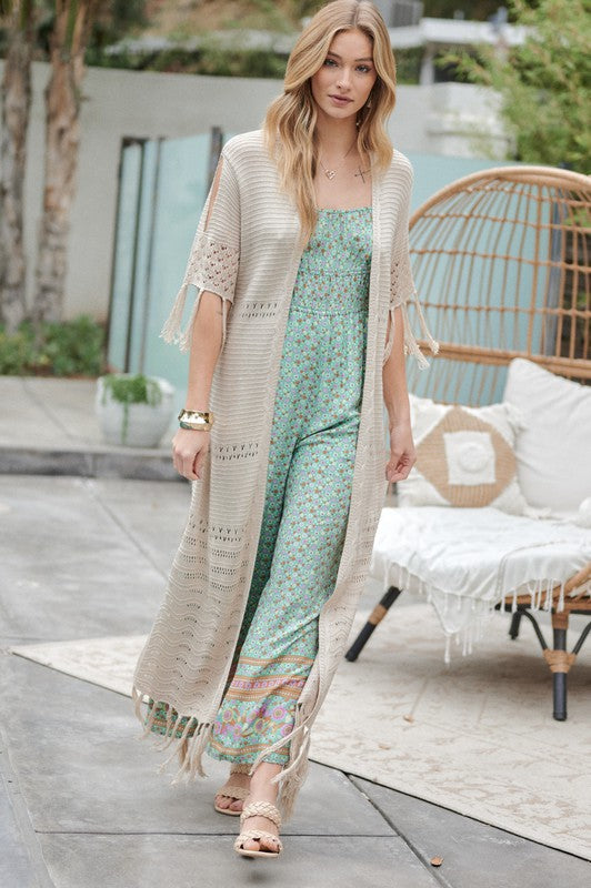 A woman in a green floral dress and a Solid Long Cardigan With Fringe exudes boho-chic vibes as she strolls on a patio, surrounded by outdoor furniture and lush plants.