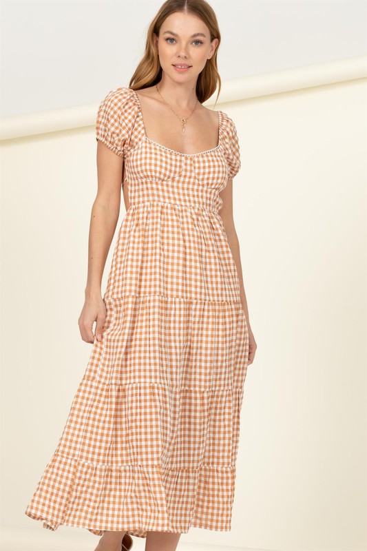 Wearing the "Somewhere to Go Tie-Back Gingham Print Maxi Dress," which features a black and white gingham print with puffed sleeves, a tiered skirt, and elegant tie-back detail, a person poses stylishly paired with black sandals.