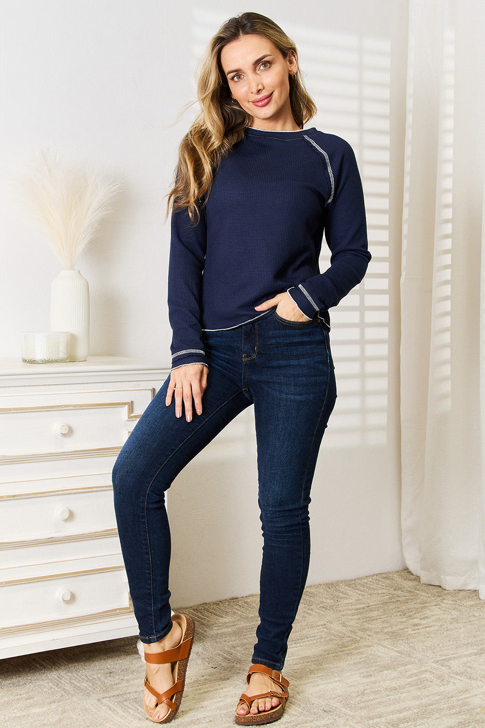 Person with long braided hair wearing a Basic Bae Long Raglan Sleeve Round Neck Top in navy blue and denim jeans, standing and smiling indoors.