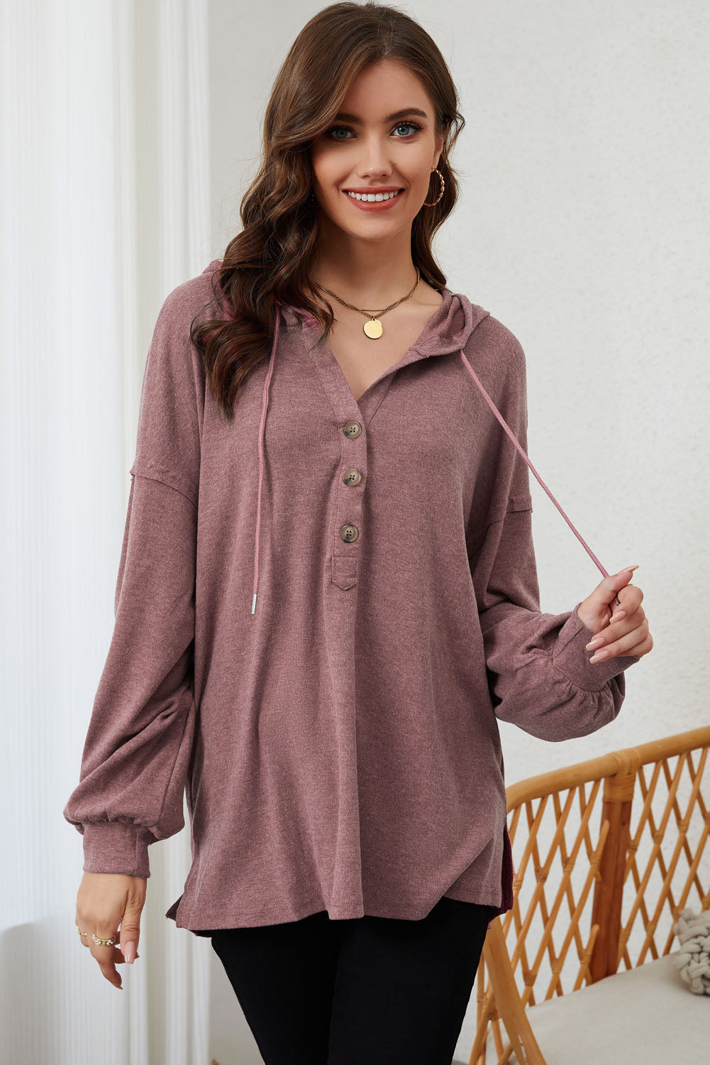 A woman stands facing away, wearing a stylish Buttoned High and Low Hem Hoodie in mauve made of high-quality fabric and black pants. She is in a room with light curtains and a wicker chair.