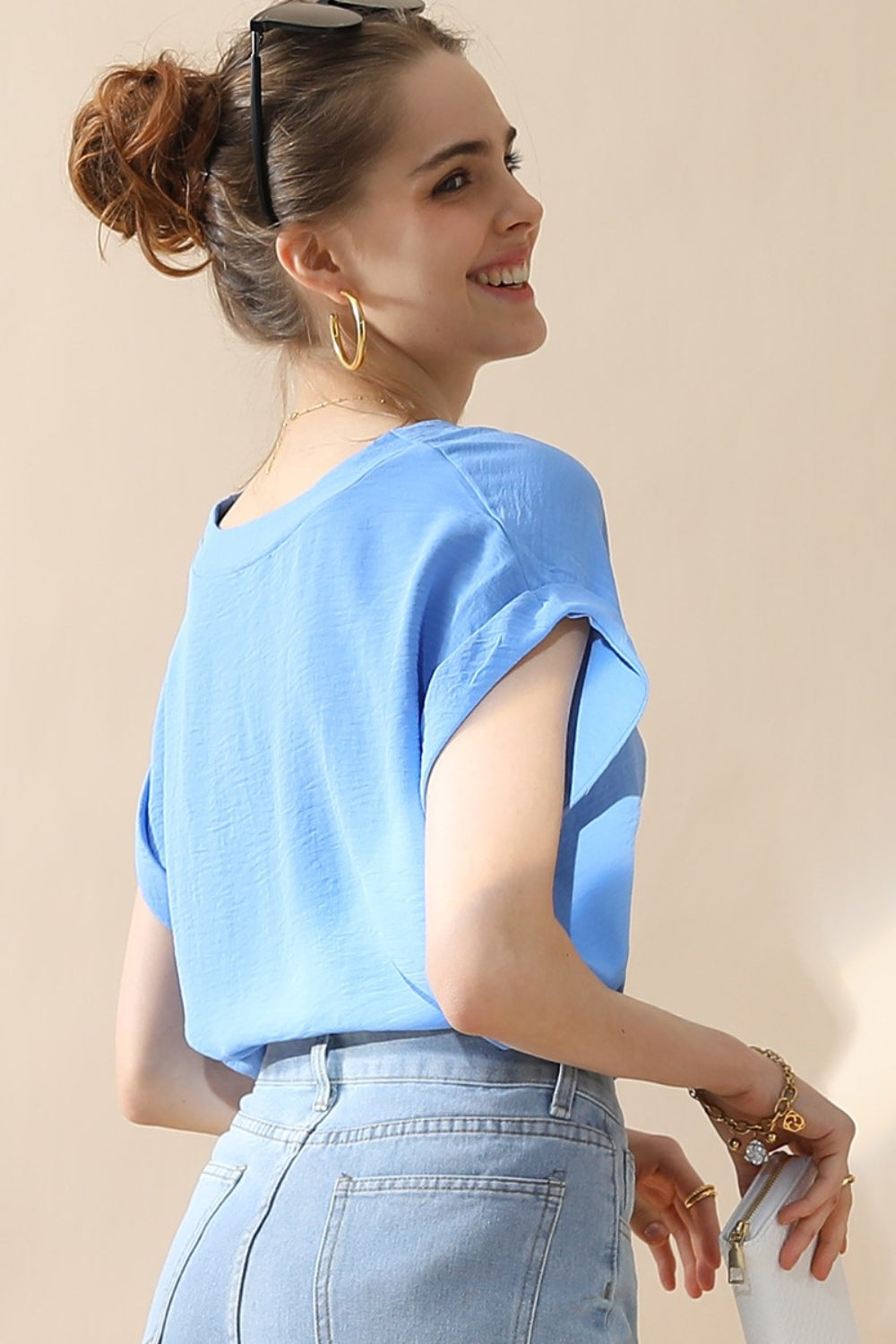 A woman wearing a stylish Ninexis V-Neck Trim Rolled Short Sleeve Shirt in light blue and high-waisted blue jeans, accessorized with hoop earrings, a necklace, and sunglasses on her head, poses in front of a beige background.