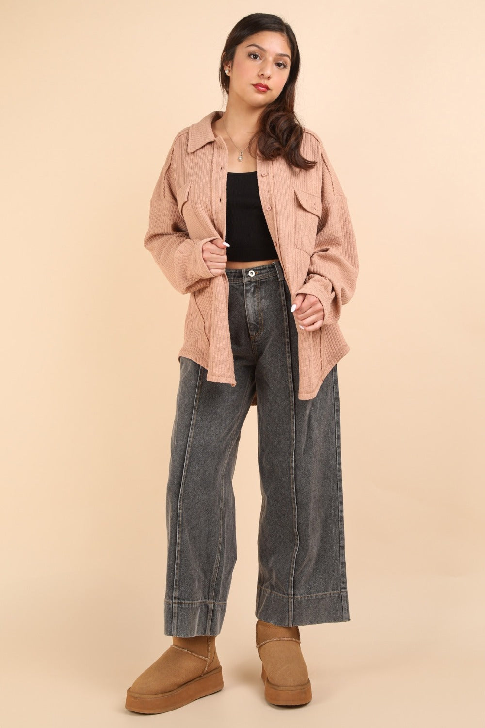 A woman wearing the VERY J Button Down Textured Knit Shacket in an oversized beige style over a black top stands against a plain background, showcasing the perfect transitional piece for any wardrobe.