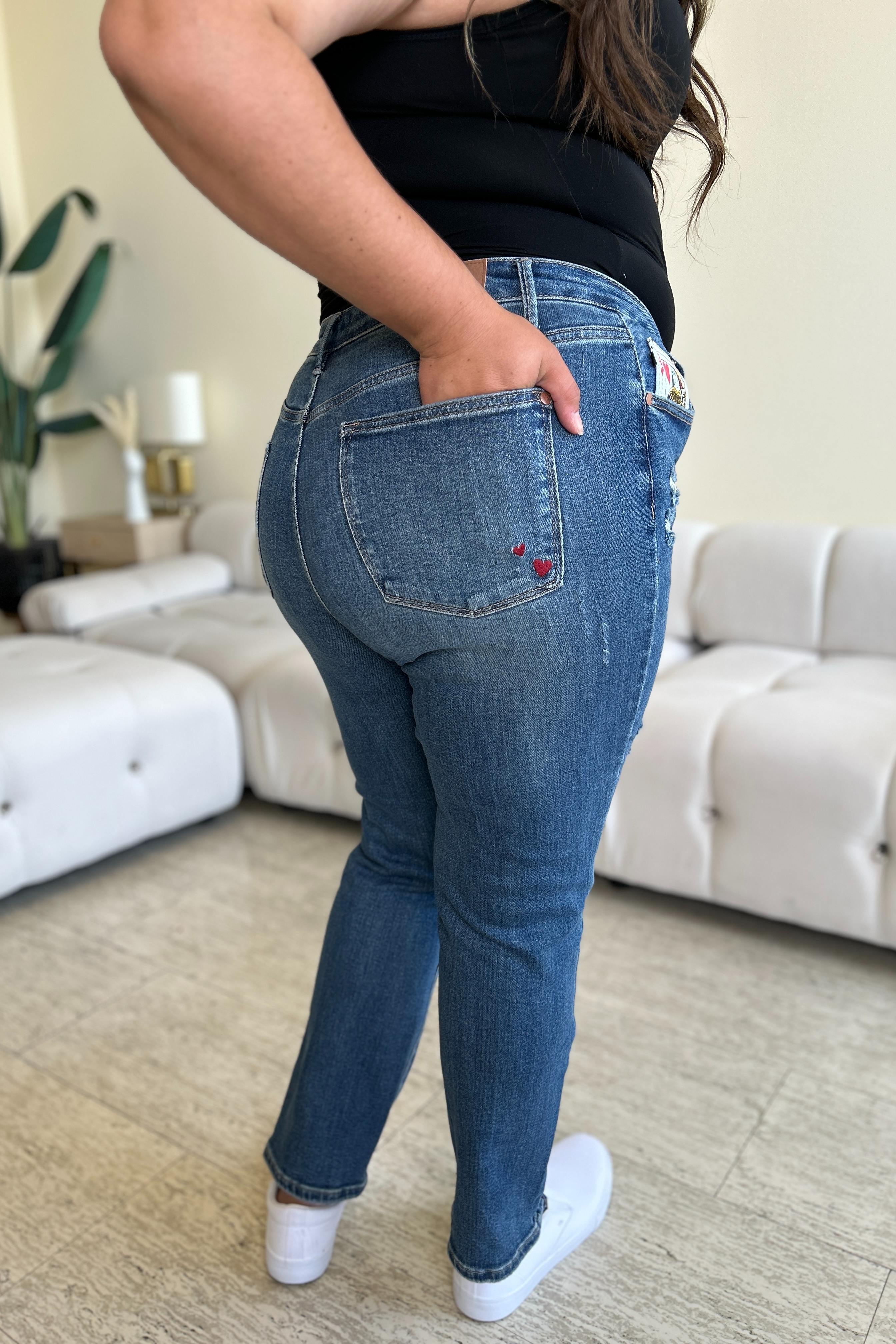 Two women stand side by side in a living room, both wearing stylish jeans and tank tops. One woman is pointing at the distinctive back pocket of her Judy Blue Full Size Queen Of Hearts Coin Pocket BF Jeans, while the other smiles.