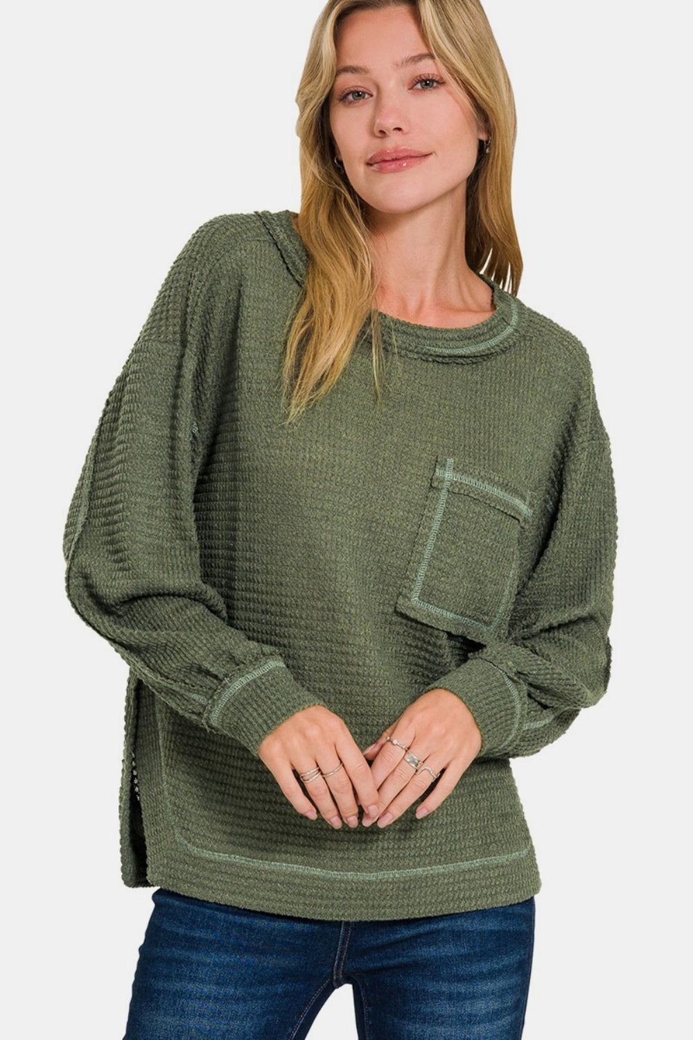 A person wearing the Zenana Exposed Seam Side Slit Long Sleeve Top in textured green, paired with blue jeans, stands against a plain background.