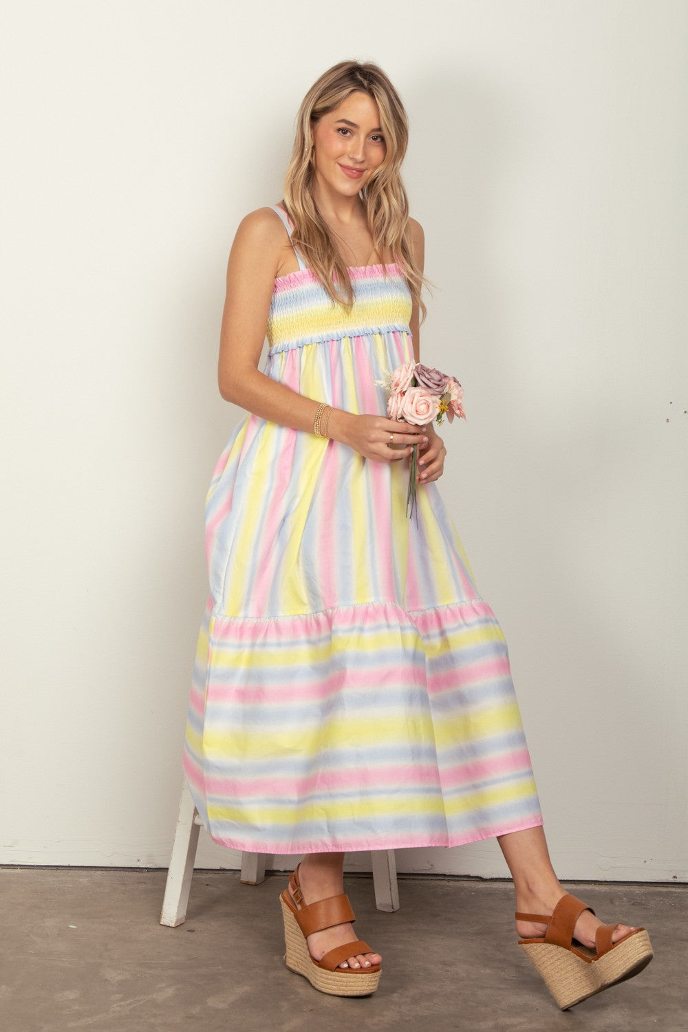 A woman in the VERY J Striped Woven Smocked Midi Cami Dress, featuring pastel stripes, holds a small bouquet of flowers. She pairs the dress with tan platform sandals that complete her chic summer look and stands against a plain white background.