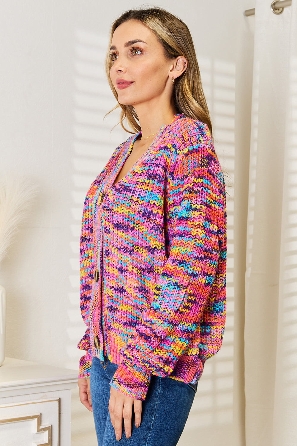 Person wearing an Angel Wings Woven Right V-Neck Long Sleeve Cardigan, characterized by its multicolored design and large buttons, paired with blue jeans. The cardigan, made of 100% acrylic knit, offers a versatile look perfect for any occasion. They are standing indoors against a white background.