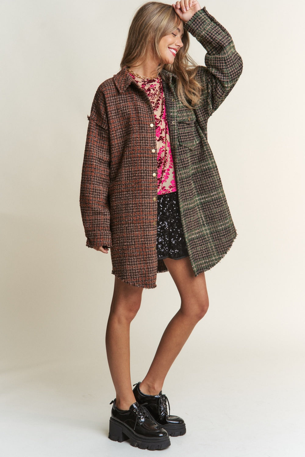 A woman wearing an eclectic mix poses against a plain background, showcasing an ideal fall wardrobe ensemble. She dons a mixed-patterned oversized jacket over a red floral sheer top and black sequined shorts. The J.NNA Snap Down Tweed Plaid Contrast Longline Shacket adds texture and depth to her look.