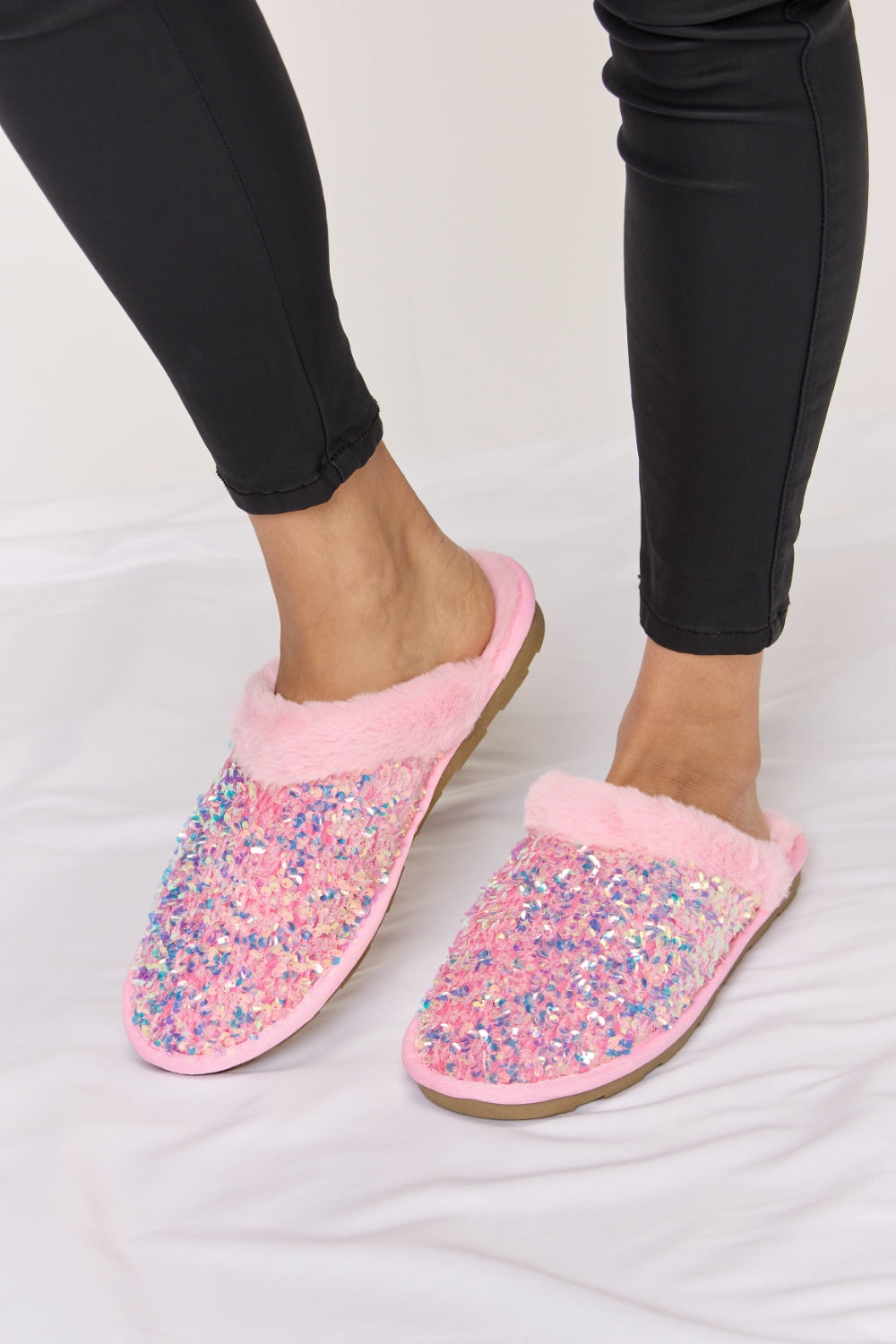 Person wearing cozy and luxurious Forever Link Sequin Plush Round Toe Slippers with fuzzy trim and black pants, sitting on a white surface.