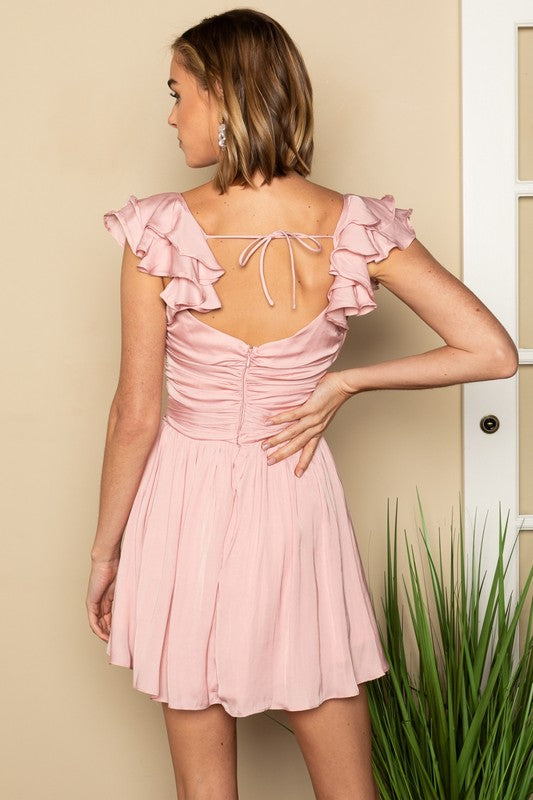 A woman wearing a stylish and playful Ruffled Surplice Mini Dress with ruffled sleeves poses beside a green plant against a beige backdrop.