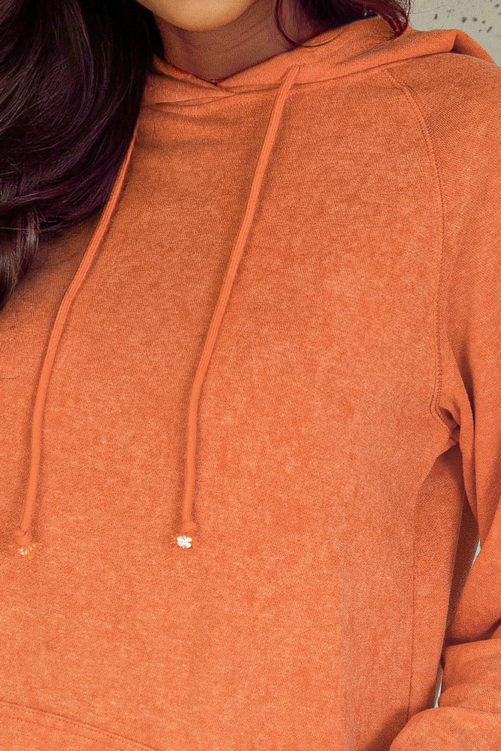 A person wearing an Orange Mineral Wash Kangaroo Pocket Drawstring Pullover Hoodie and sunglasses smiles while looking over their shoulder against a speckled wall backdrop.