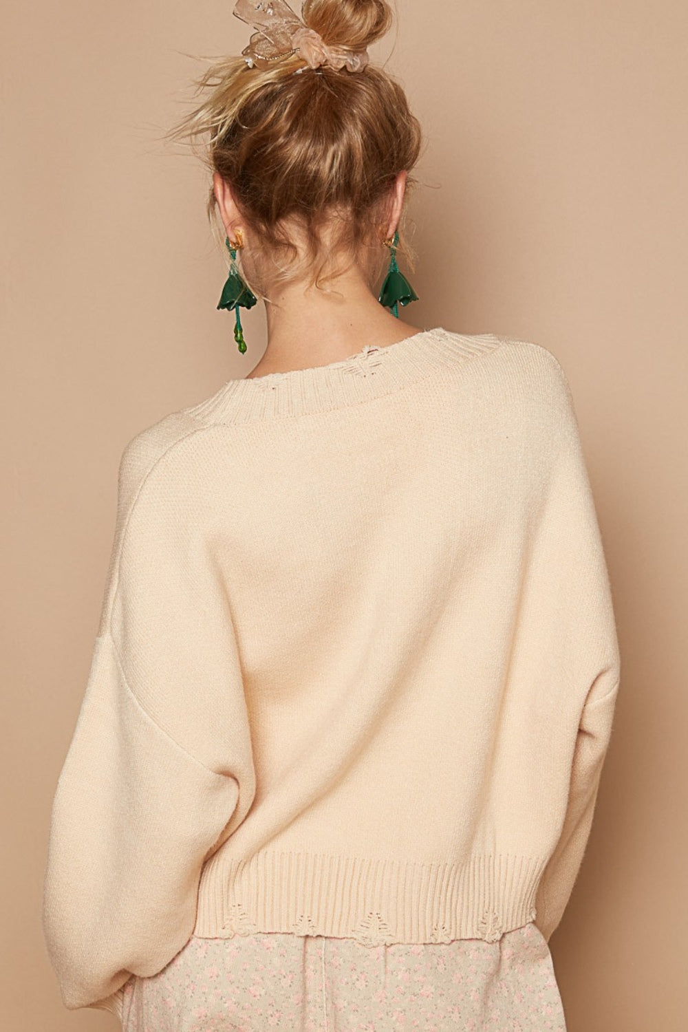 A woman smiles, donning the POL Round Neck Long Sleeve Peace Sign Sweater in beige, green tassel earrings, and beige patterned pants.