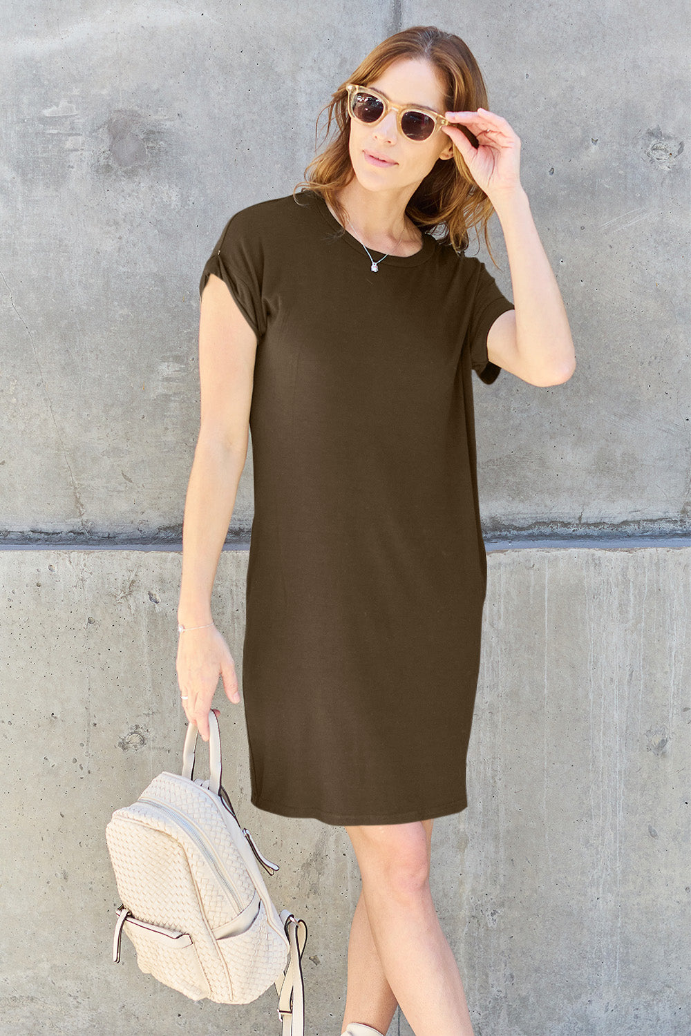 A woman wearing a dark olive-green Basic Bae Full Size Round Neck Short Sleeve Dress with Pockets and sunglasses stands against a concrete wall, holding a light-colored backpack.