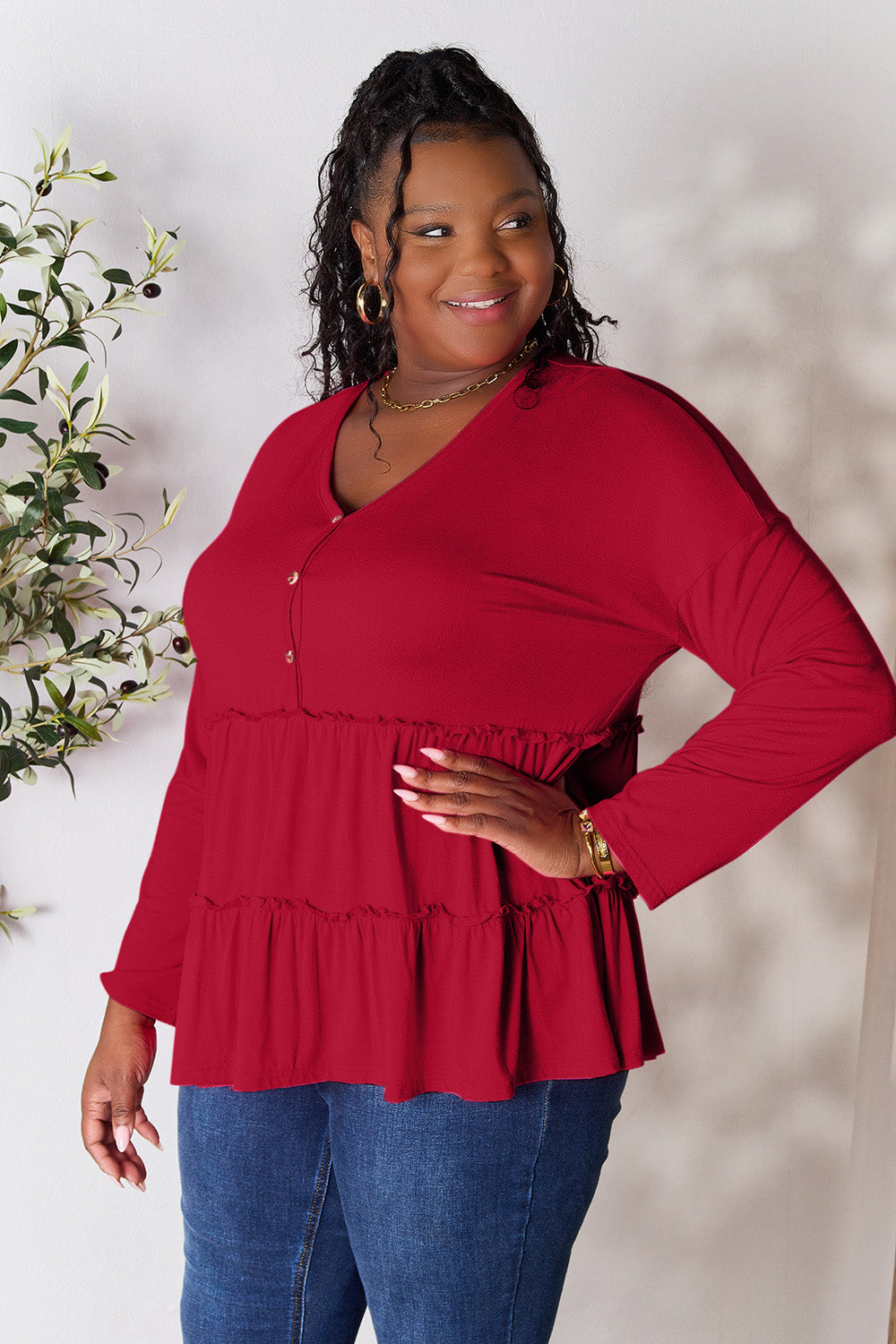 Person stands indoors near a plant, wearing the Double Take Half Button Long Sleeve Ruffle Hem Blouse in gray and blue jeans, smiling while looking to the side. The slightly stretchy fabric of the blouse adds comfort while its buttoned design offers a touch of elegance.