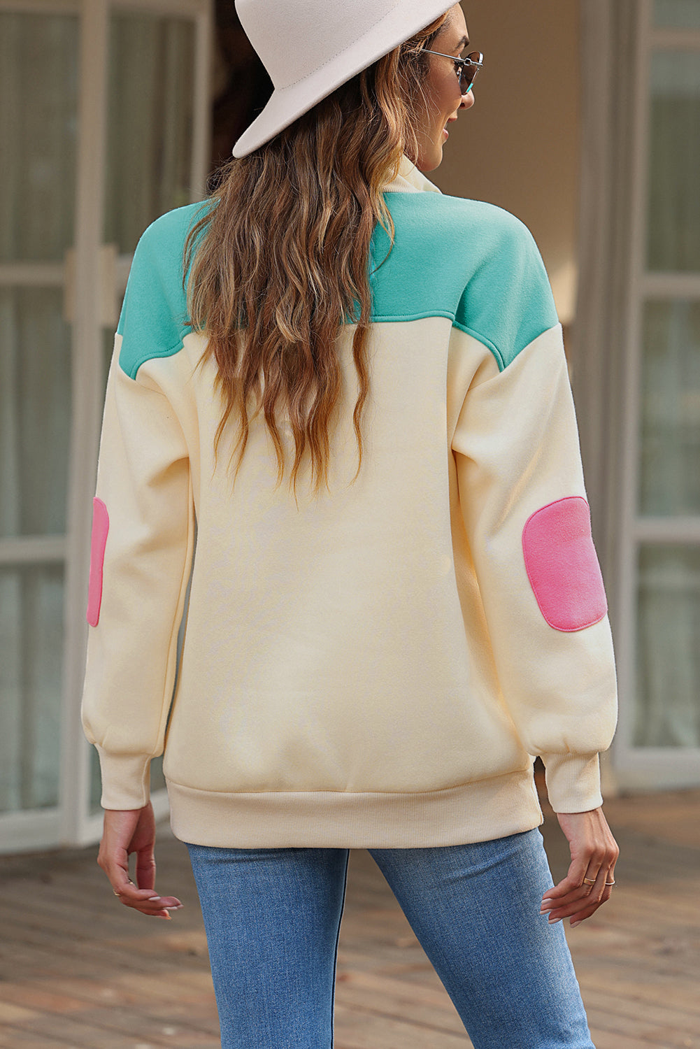 A woman wearing the Apricot Color Block Elbow Patch Half Button Sweatshirt, featuring cream and mint colors with pink elbow patches and a turn-down collar with snap button closure, stands outdoors. She has long wavy hair, sunglasses, a hat, and is wearing blue jeans.