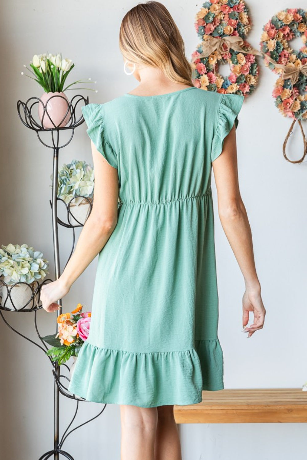 A person in a Heimish Full Size Short Sleeve V Neck Ruffled Hem Dress stands indoors, elegantly holding a hat. The floral decor in the background complements the dress's feminine charm.