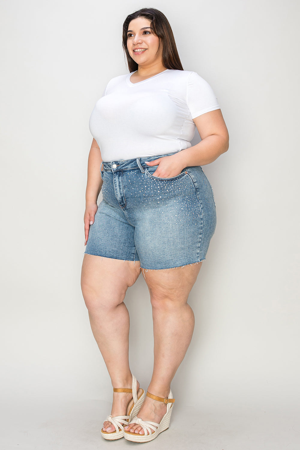 A woman wearing a white T-shirt, Judy Blue Full Size High Waist Rhinestone Decor Denim Shorts, and white sandals poses against a plain background.