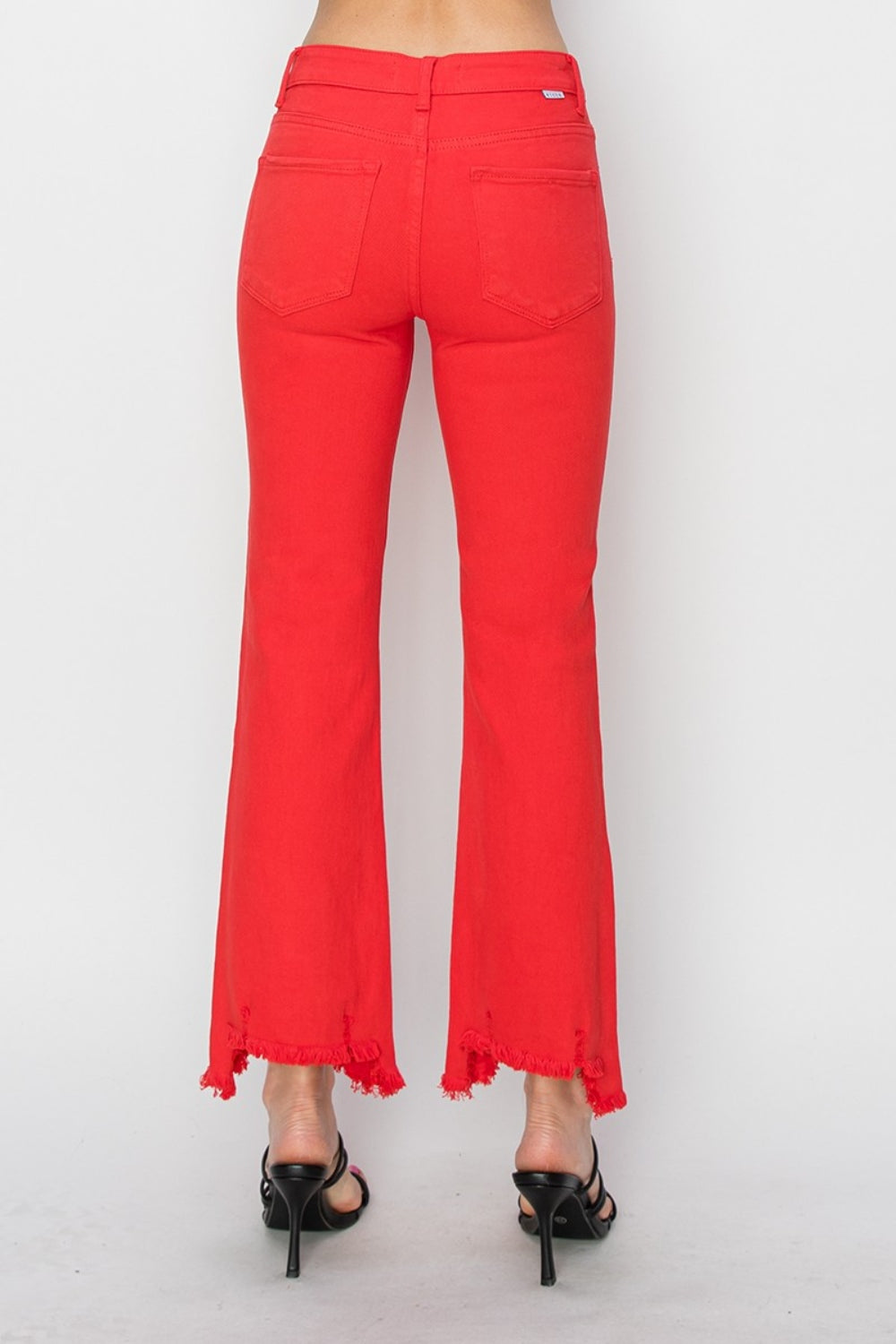 A person is wearing the RISEN Raw Hem Bootcut Jeans with Pockets in bright red, paired with a white top and black strappy high-heeled sandals. The image focuses on the stylish and functional lower half of the body.