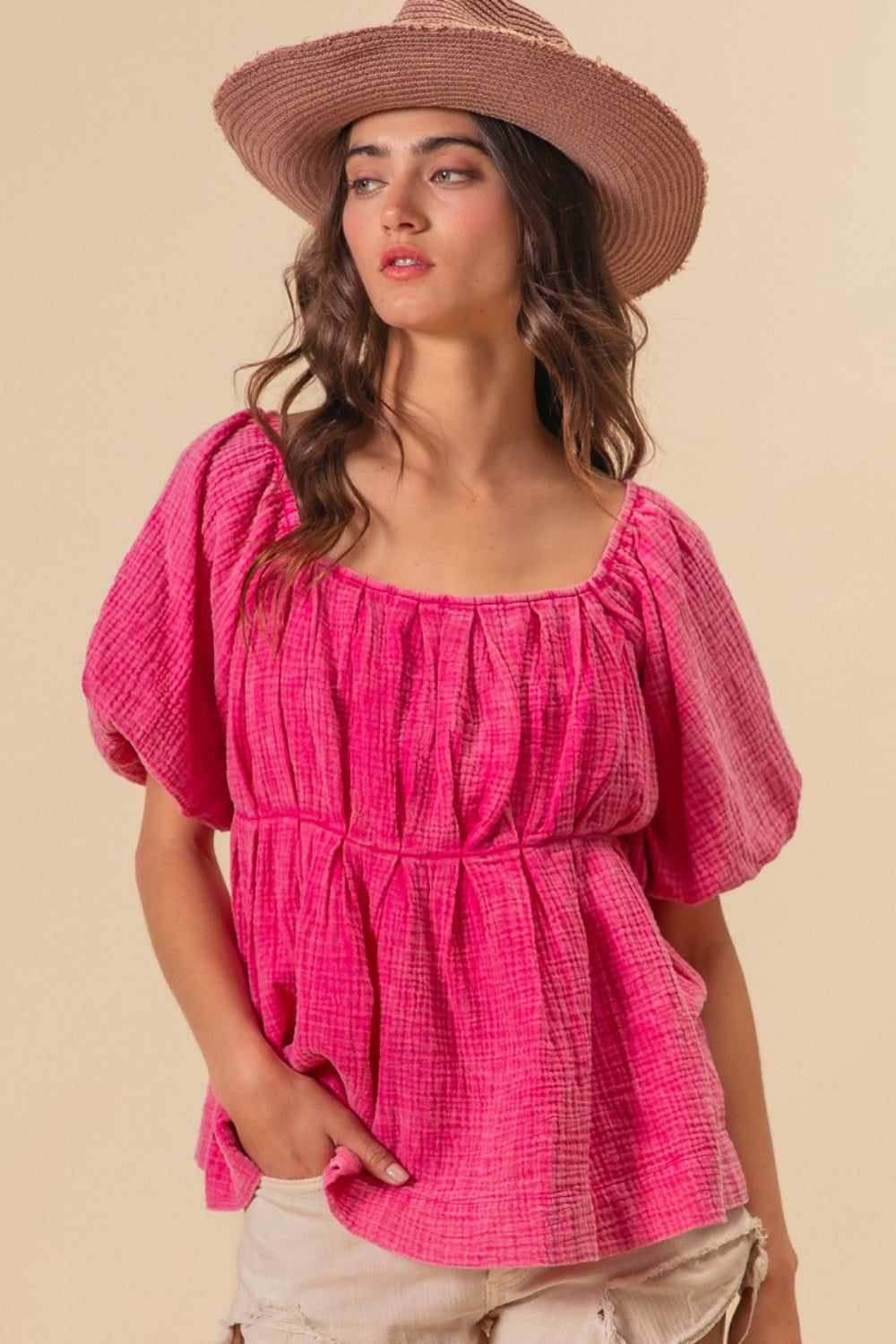 A woman wearing the BiBi Pleated Puff Sleeve Washed Blouse in pink, along with a wide-brimmed hat, strikes a pose against a beige background with one hand resting in her pocket.