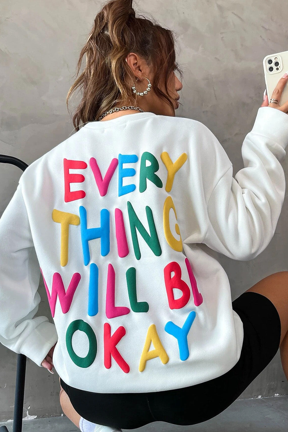 A person wearing a white beanie, the EVERY THING WILL BE OKAY Colorful Letters Sweatshirt, and sunglasses, holding a coffee cup, stands outside near windows.