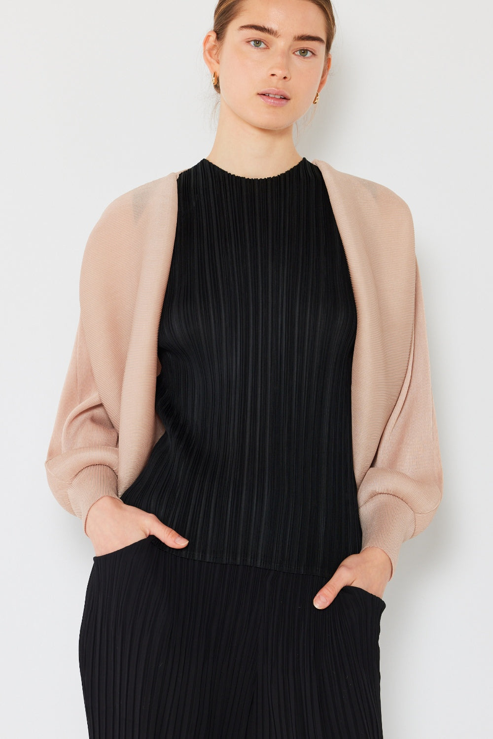 A person stands wearing the stylish Marina West Swim Rib Pleated Puff Sleeve Bolero Cardigan in beige, featuring draped sleeves, paired with a trendy black pleated top.