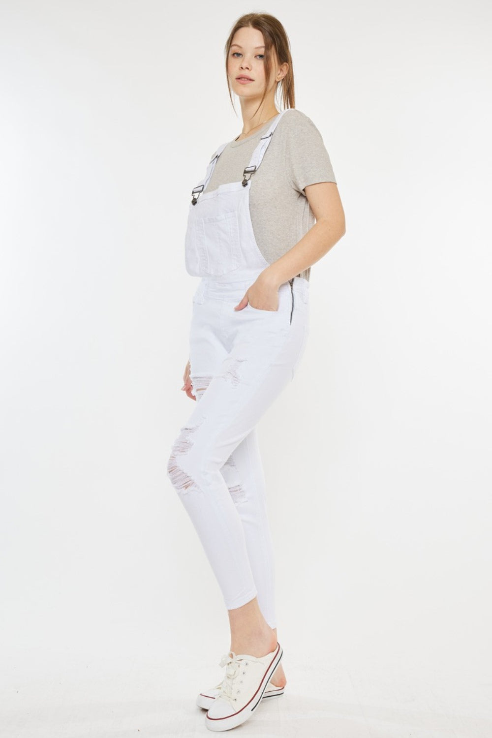 A fashion-forward woman stands against a plain background, wearing a grey t-shirt, Kancan Distressed Skinny Denim Overalls in white, and white sneakers with red detailing.