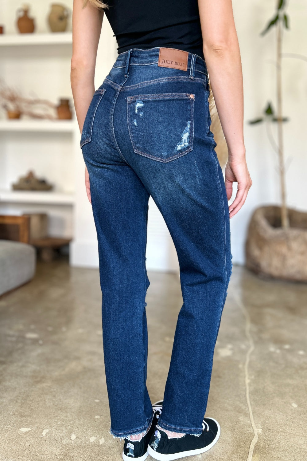 A woman in a black sleeveless top and Judy Blue Full Size High Waist Rigid Magic Heavy Destroy Straight Jeans stands indoors with her hands in her pockets. The background features a plant and shelves adorned with decor items.