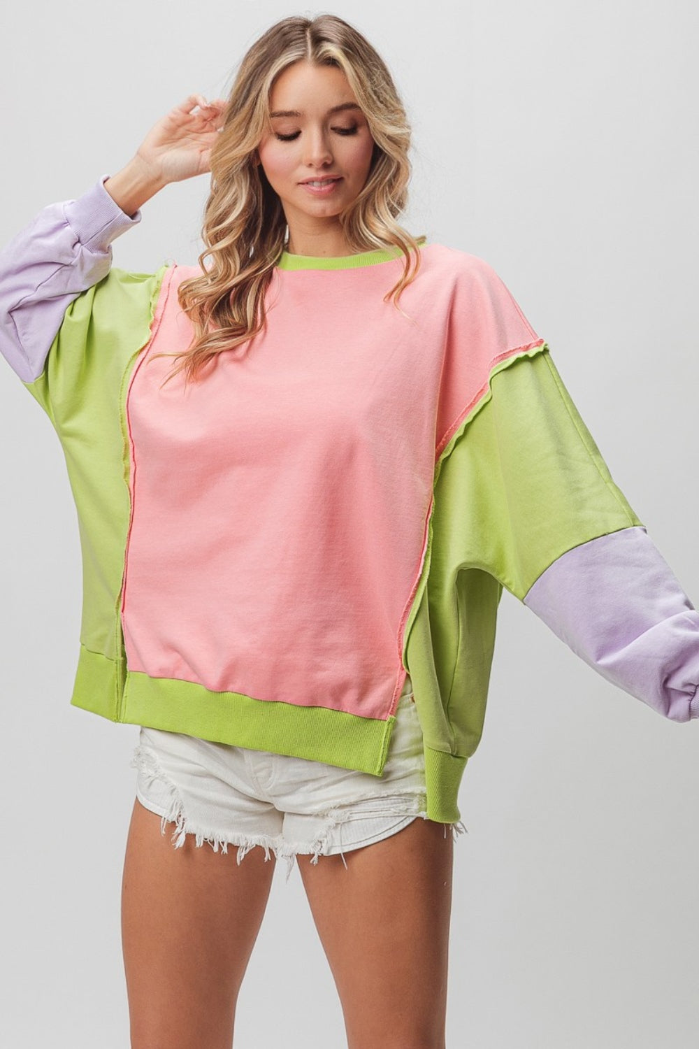 A woman is wearing a BiBi Washed Color Block Sweatshirt featuring vintage-inspired sections in pink, green, and purple. She has paired it with white distressed shorts and is posing with one arm slightly raised, perfectly showcasing the relaxed fit.