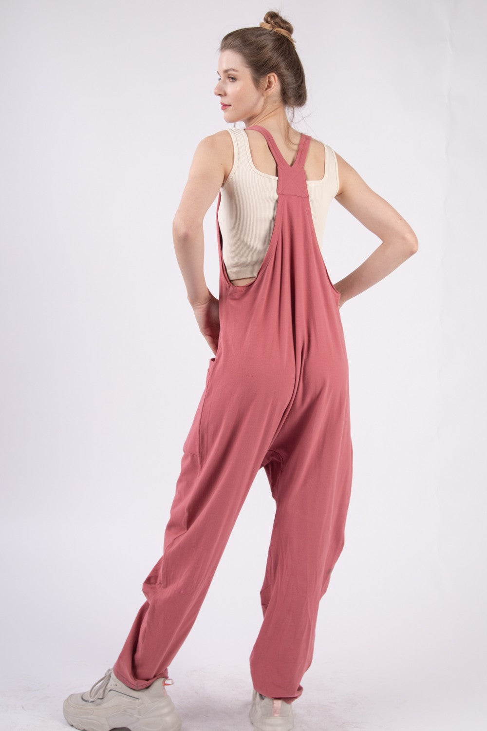 Someone is standing against a plain backdrop, wearing the VERY J Plunge Sleeveless Jumpsuit in pink and white shoes, with their hands casually tucked into the jumpsuit's pockets.