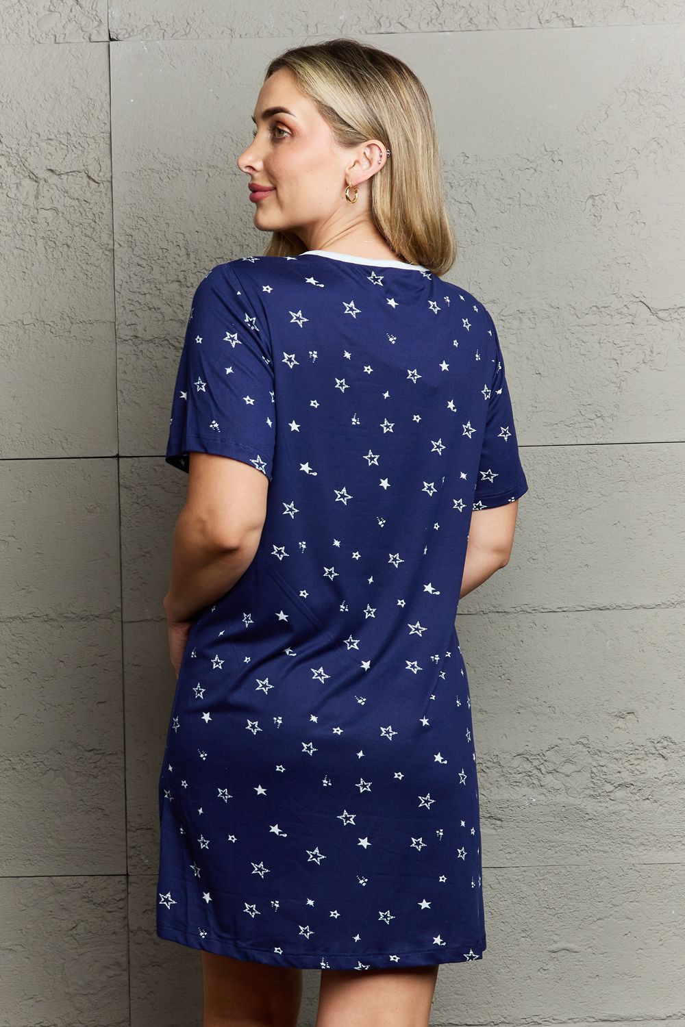 A woman with long blonde hair is wearing the MOON NITE Quilted Quivers Button Down Sleepwear Dress, which is navy blue and adorned with white stars; she stands against a textured gray wall.