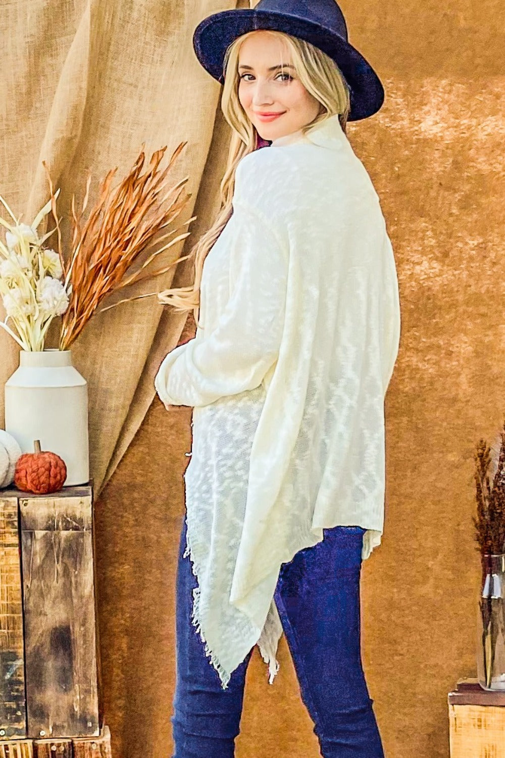 A person with long blonde hair wears a black hat, the And The Why Textured Knit Fringe Hem Cardigan in white, and blue jeans, standing against a brown backdrop near a wooden table with a vase of dried flowers and a small pumpkin.