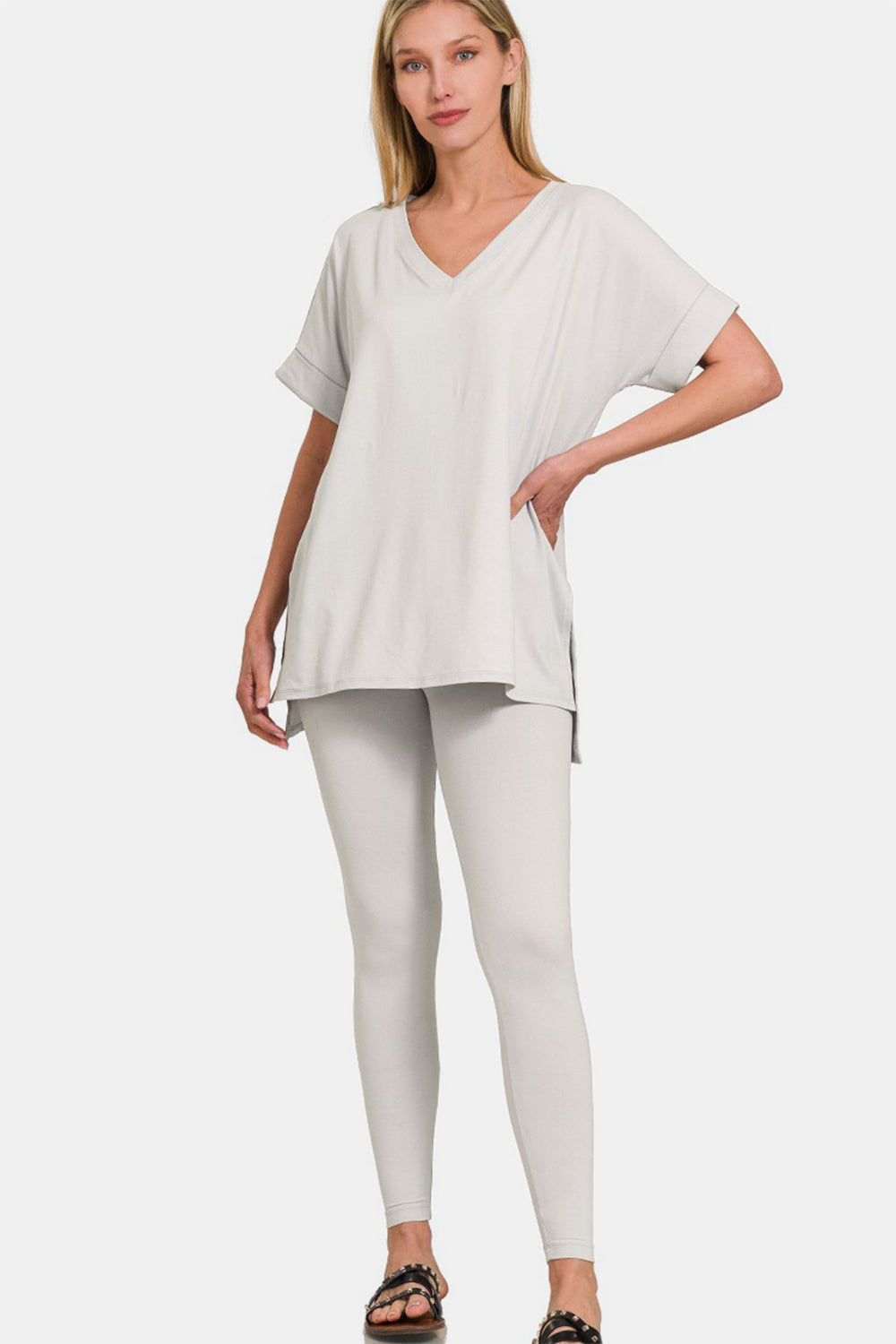 The individual gracefully dons the Zenana V-Neck Rolled Short Sleeve T-Shirt and Leggings Lounge Set in white, standing confidently with one hand on their hip. Paired with sandals, this ensemble captures both comfort and style against a simple backdrop.