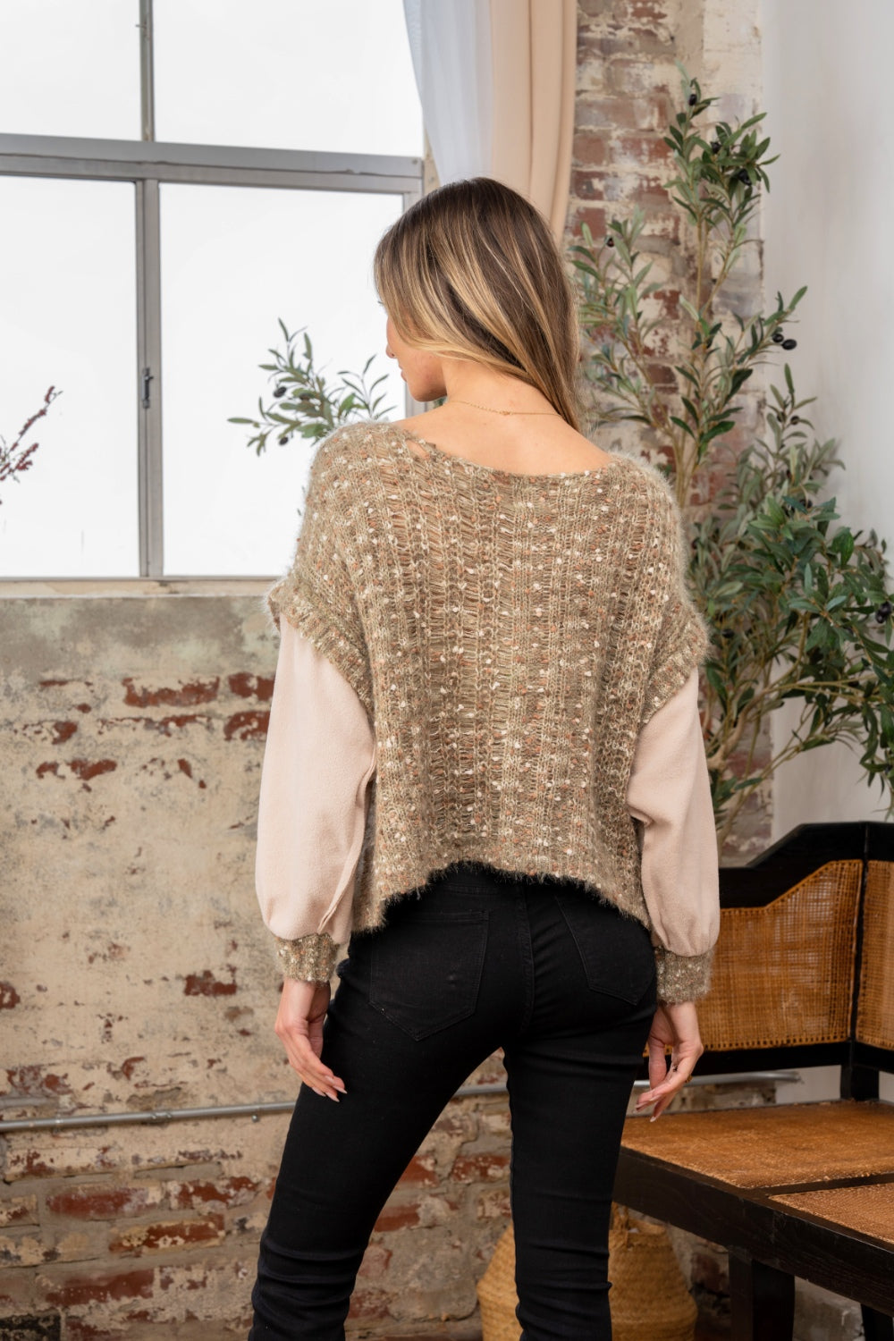 Standing indoors by a window, a person looks cozy and stylish in black pants, their textured sweater contrasting perfectly with the brick wall and greenery backdrop. The Sew In Love Full Size Fuzzy Long Sleeve Knit Top adds an elegant touch as one hand rests in their pocket.