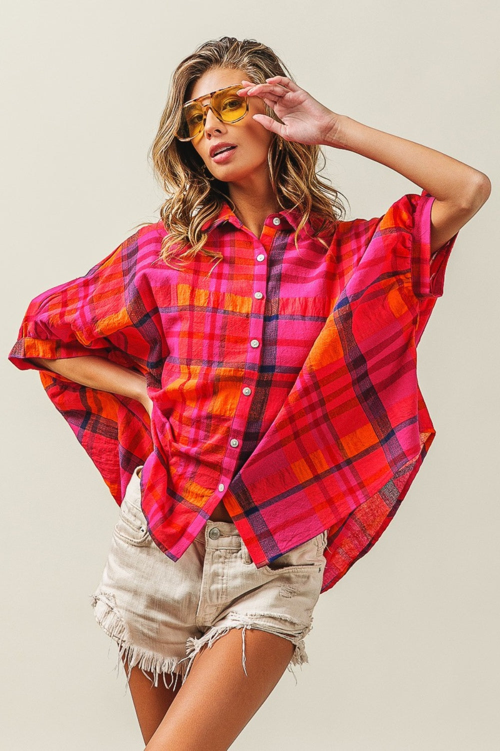 A woman outfitted in a BiBi Button Up Dolman Sleeve Plaid Shirt, paired with a white crop top, distressed denim shorts, and yellow sunglasses.