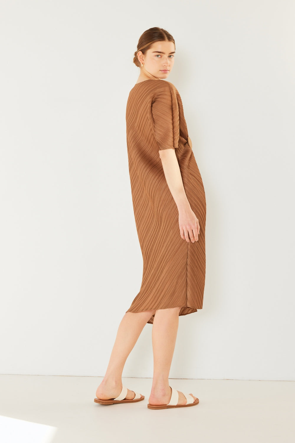 A woman stands against a plain backdrop wearing the stylish Marina West Swim Pleated Dolman Sleeve Dress, which features a knee-length, short-sleeved design in a textured brown fabric with an eye-catching circular detail at the waist. She completes her look with white sandals. This modern chic dress exudes versatility, making it perfect for various occasions.