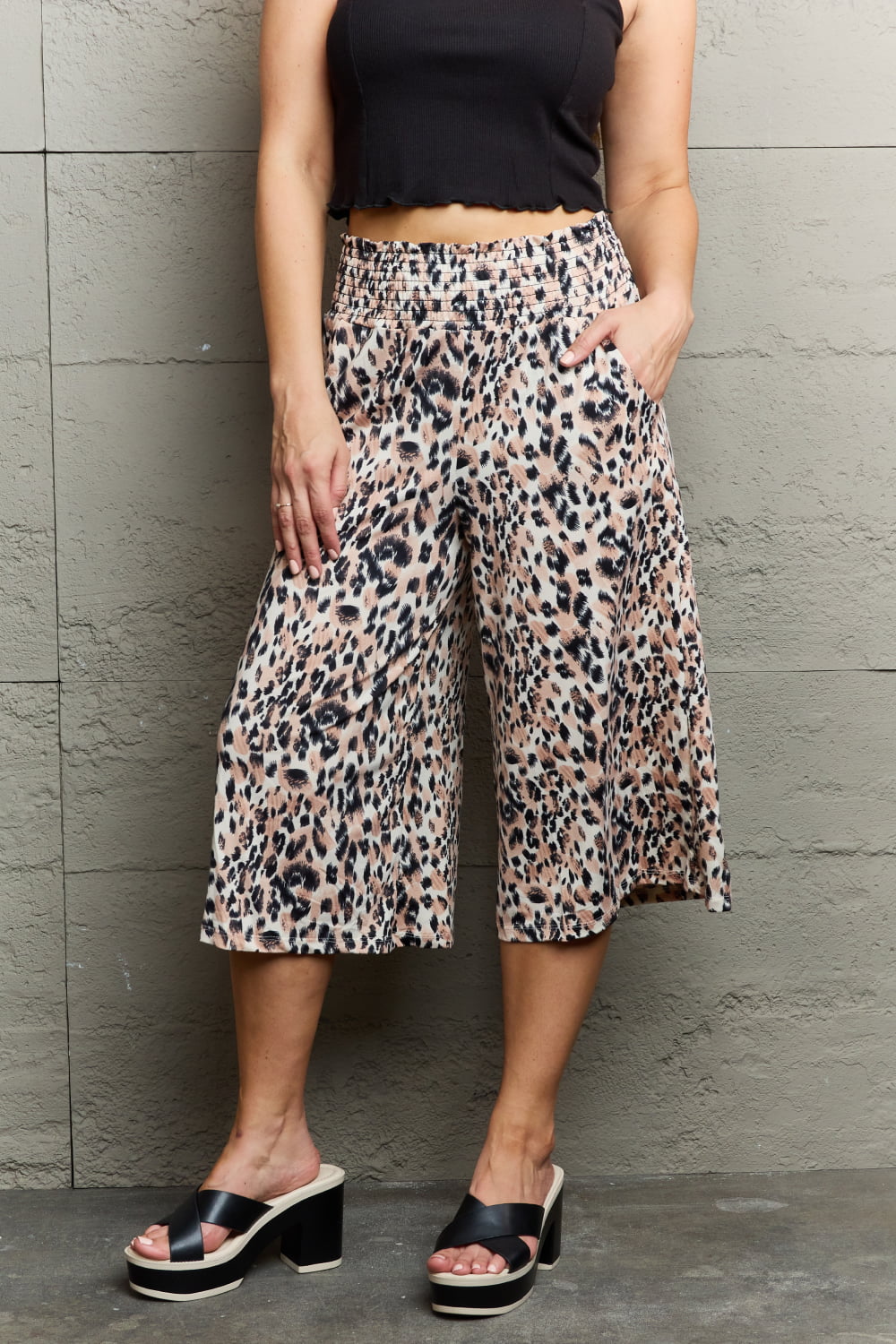 A person standing against a wall, wearing a black sleeveless top, chic Ninexis Leopard High Waist Flowy Wide Leg Pants with Pockets, and black platform sandals. One hand is in their pocket.