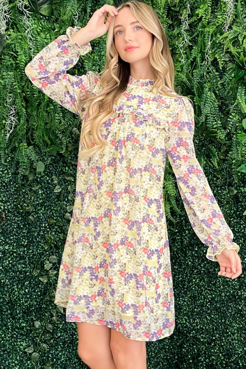 A woman stands in front of a leafy green background wearing the And The Why Floral Mock Neck Flounce Sleeve Dress, smiling and looking to the side.