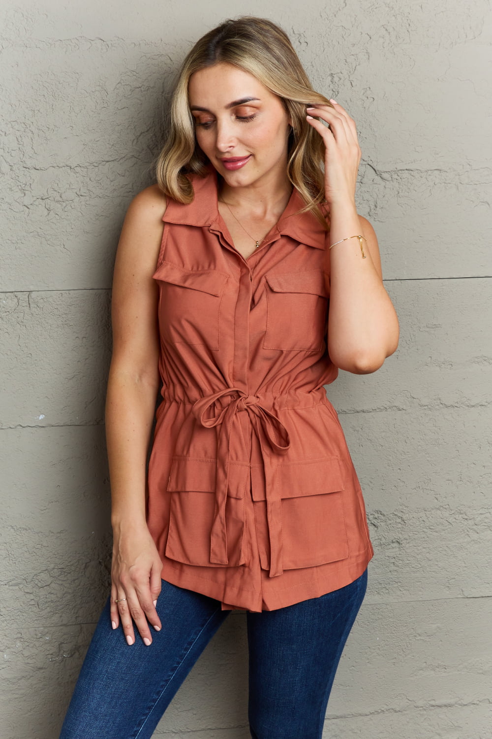 A woman wearing the Ninexis Follow The Light Sleeveless Collared Button Down Top in rust-orange with a tied waist poses in front of a textured grey wall, touching her hair while looking down. She pairs the top with blue jeans featuring an elastic waistband.