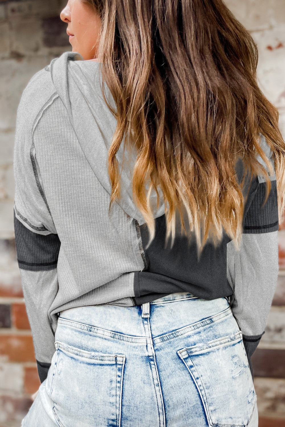 Rear view of a person with long hair wearing a stylish Gray Colorblock Seamed Ribbed Henley Hoodie and light-washed jeans, standing against a brick wall.