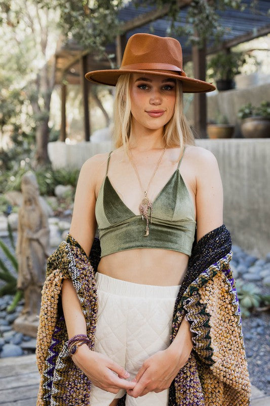 A woman with long blonde hair wears a brown hat, a green Velvet Longline Bralette with adjustable straps, a patterned shawl, and multiple bracelets, standing outdoors near plants and pottery.