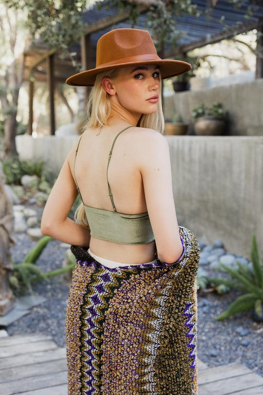 A woman with long blonde hair wears a brown hat, a green Velvet Longline Bralette with adjustable straps, a patterned shawl, and multiple bracelets, standing outdoors near plants and pottery.