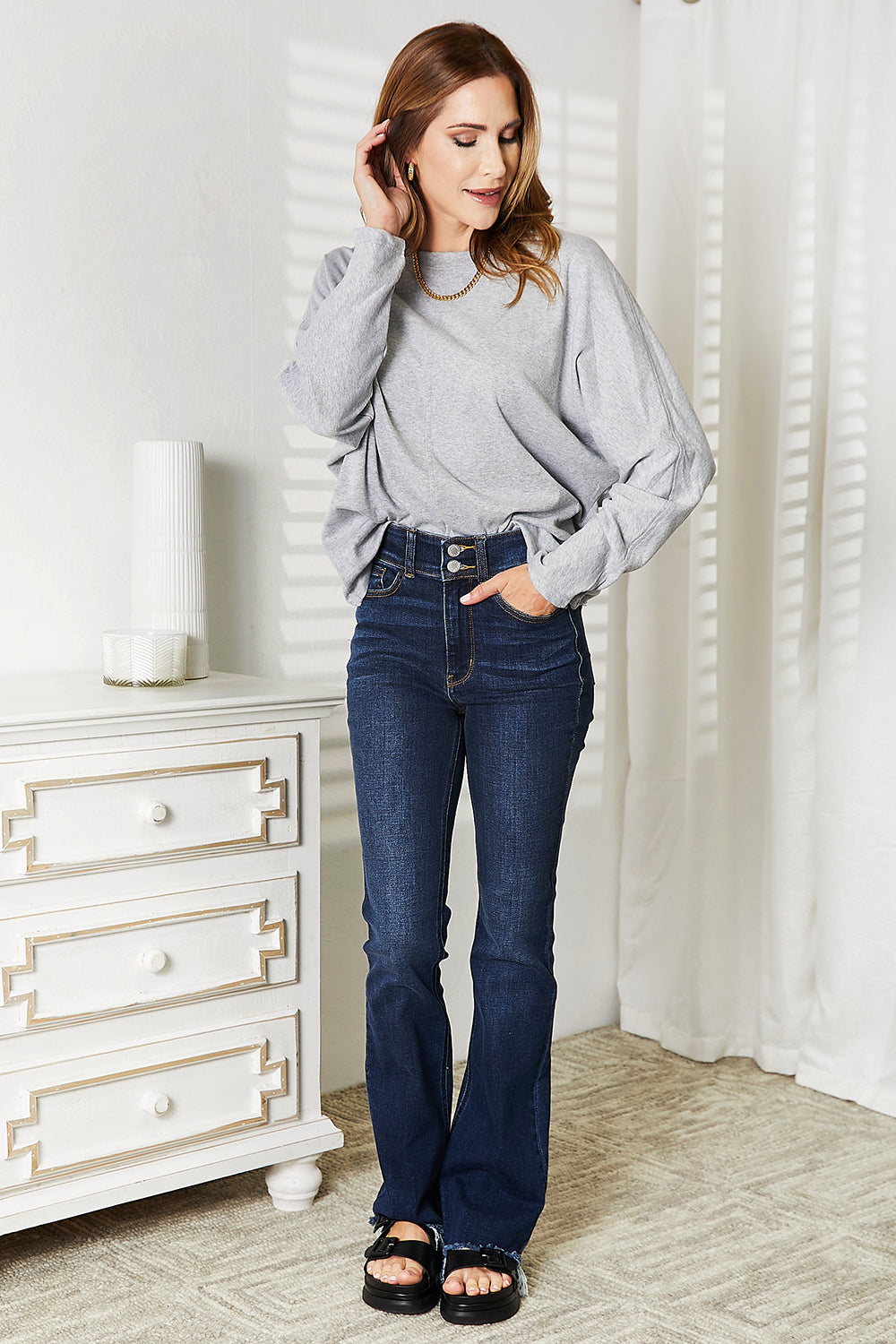 A person with long hair wearing a Double Take Seam Detail Round Neck Long Sleeve Top in solid black and blue jeans stands with one hand in their pocket, looking downward and smiling.