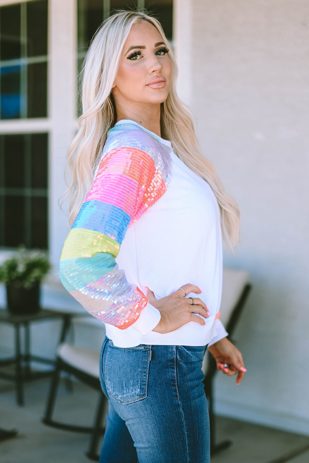 A person with long blonde hair is seen from the back, wearing a White Sequin Color Block Raglan Sleeve Pullover Sweatshirt and blue jeans. They are standing in front of a building with large windows.