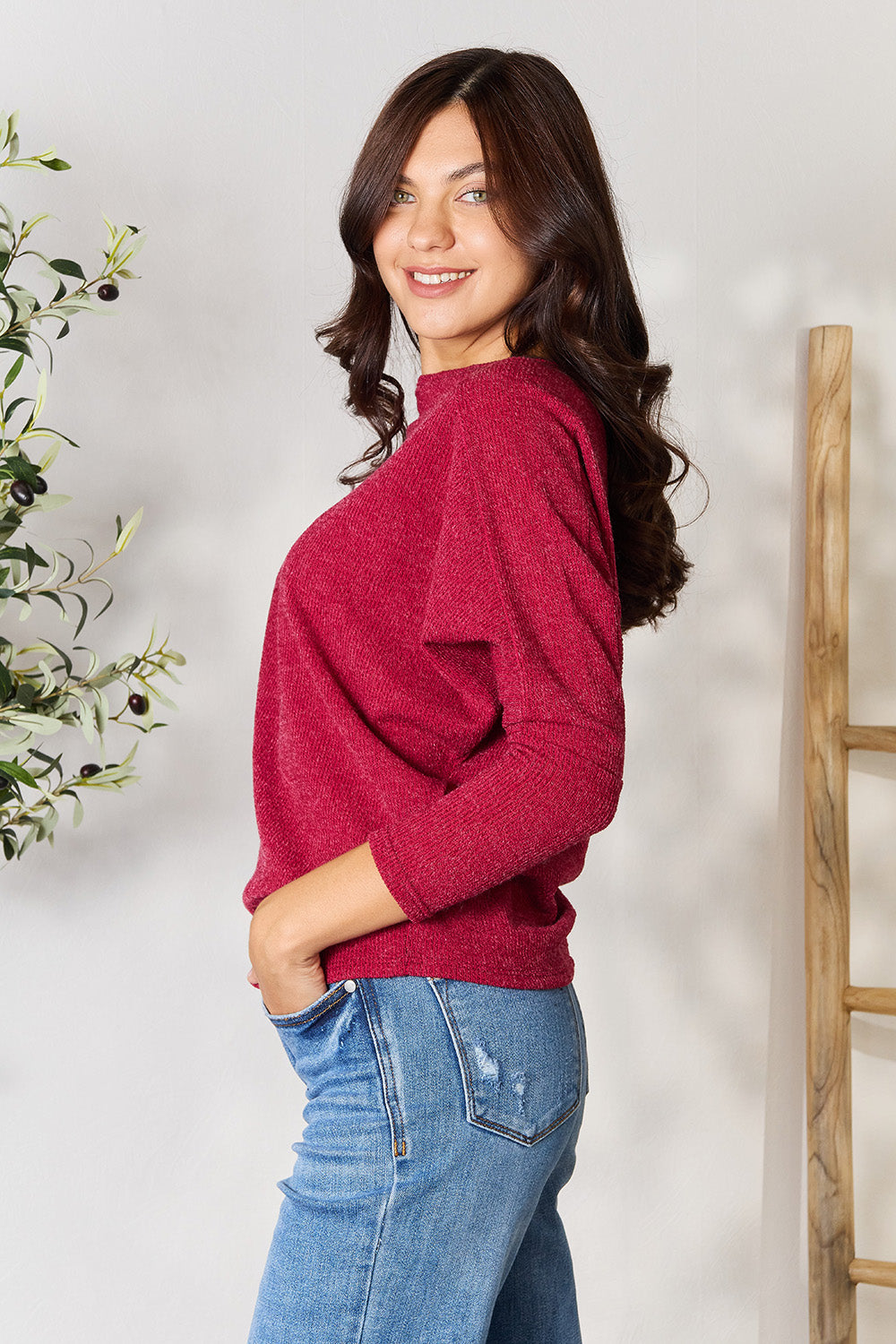 Person wearing a BOMBOM Drop Shoulder Long Sleeve Blouse in red, paired with blue jeans, standing with one hand in their pocket and the other touching their neck, in front of a white wall with a potted plant on the side.