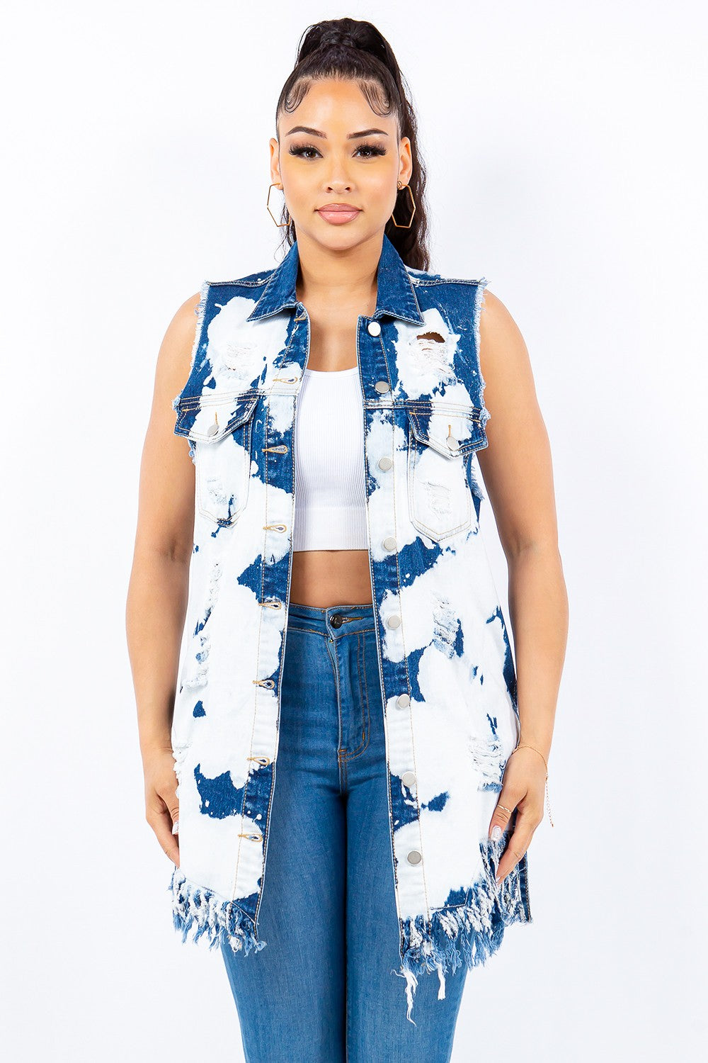 A person standing against a plain white background, wearing an American Bazi Distressed Frayed Edge Longline Denim Vest, paired with a white crop top and blue jeans. The person has their hair styled in a high ponytail and is facing the camera.