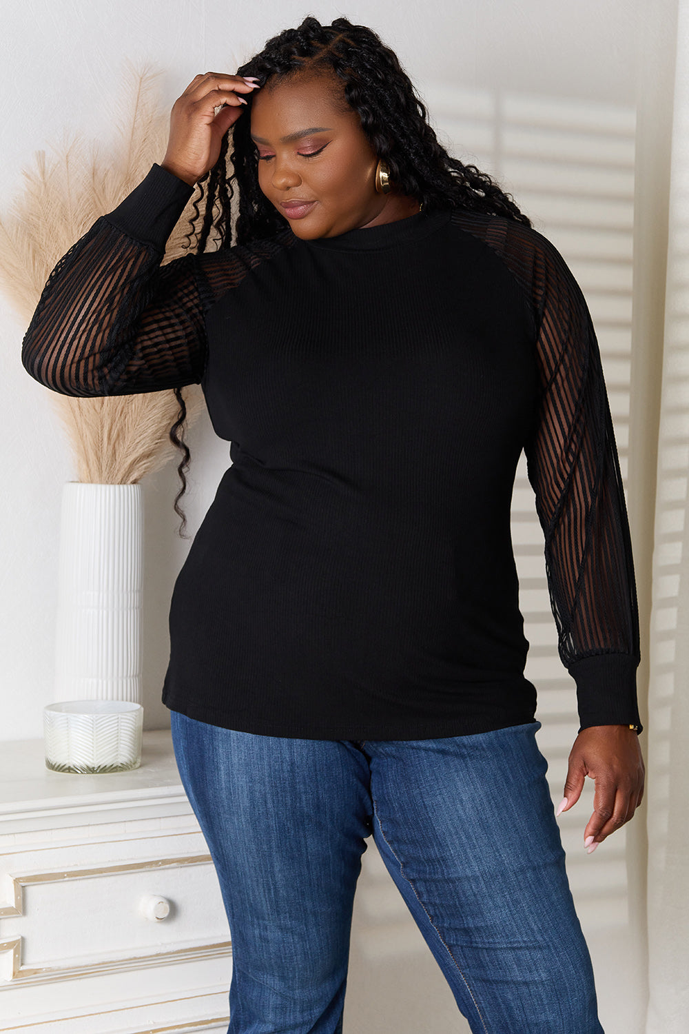 A woman with long dark hair stands indoors wearing a versatile wardrobe piece: the Double Take Round Neck Raglan Sleeve Blouse paired with blue jeans, her right hand in her pocket. A white vase with pampas grass is on the table next to her.