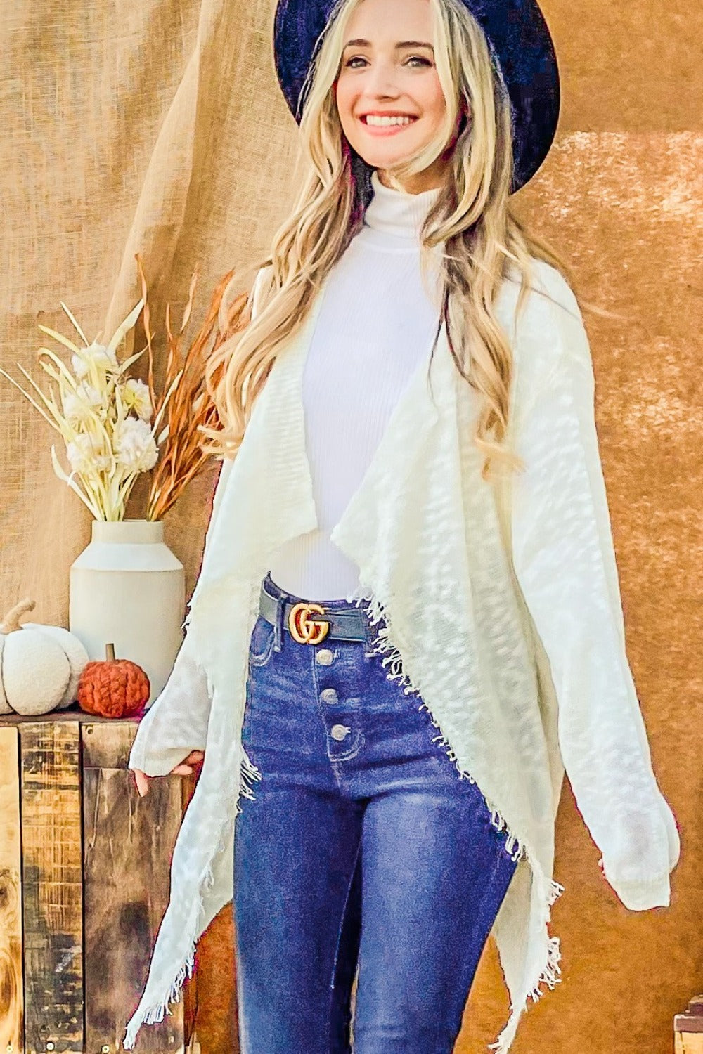 A person with long blonde hair wears a black hat, the And The Why Textured Knit Fringe Hem Cardigan in white, and blue jeans, standing against a brown backdrop near a wooden table with a vase of dried flowers and a small pumpkin.